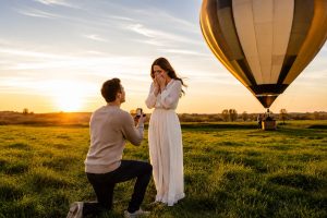 Man vraagt zijn partner ten huwelijk op één knie bij zonsondergang, met een luchtballon op de achtergrond.