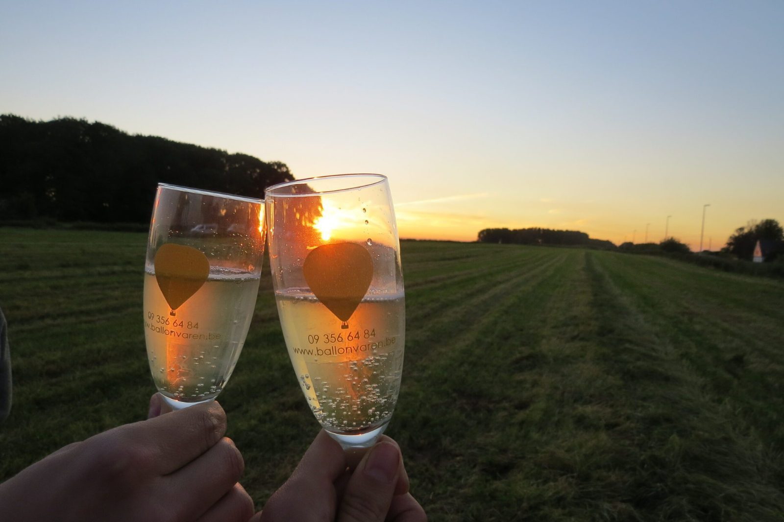 Twee glazen bubbels van ballonvaren.be klinken tegen de ondergaande zon tijdens de après-ballooning na een ballonvaart voor moederdag